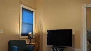 Una sala de estar con ventilador de techo, televisor de pantalla plana sobre un mueble bajo, un sillón azul y una ventana con persianas.