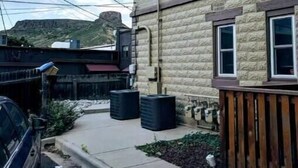 Un edificio de dos pisos con fachada de piedra, un poste de electricidad visible y un área de estacionamiento con un automóvil y contenedores de almacenamiento negros.