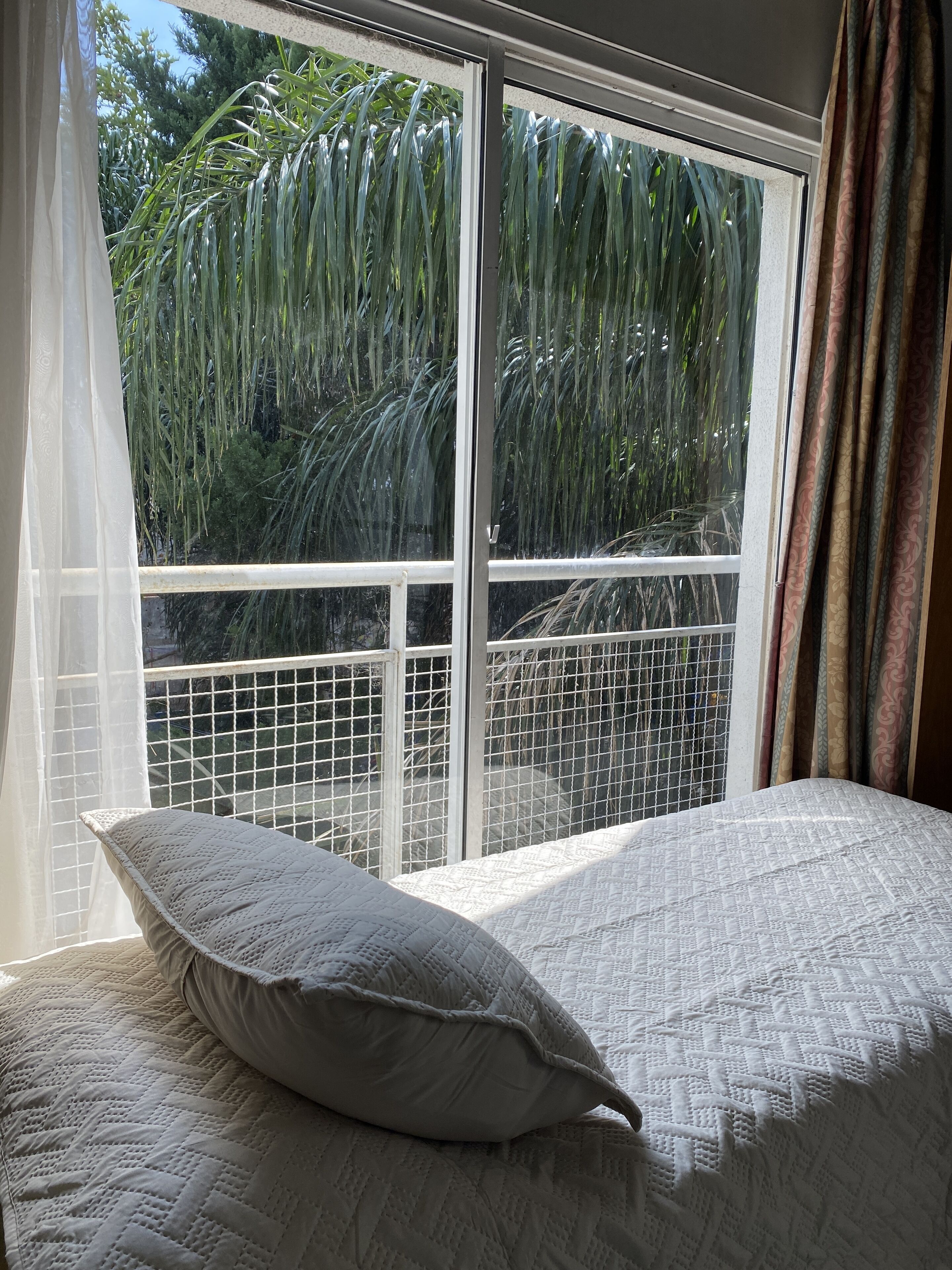 A bedroom with a bed, a window with a view of palm trees, and a balcony with a metal railing.