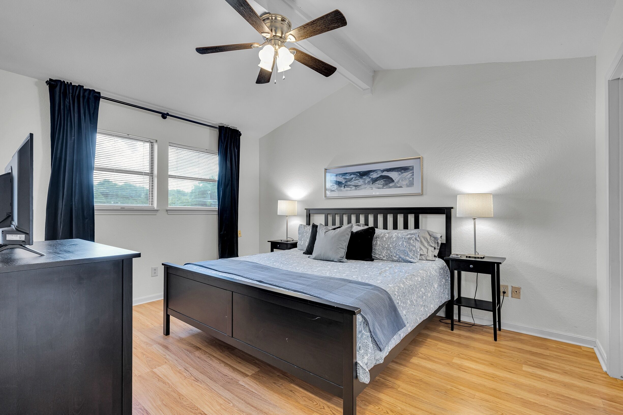 A bedroom with a bed, bedside tables, a ceiling fan, a window with curtains, and a wall-mounted picture.