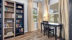 A room with a bookshelf, a table with a chair, and two windows with curtains.