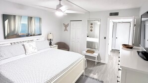 A bedroom with a bed, a desk, a chair, a mirror, a ceiling fan, and a door. - Cozy, spacious seaside condo in Ocean Village! (Fort Pierce)