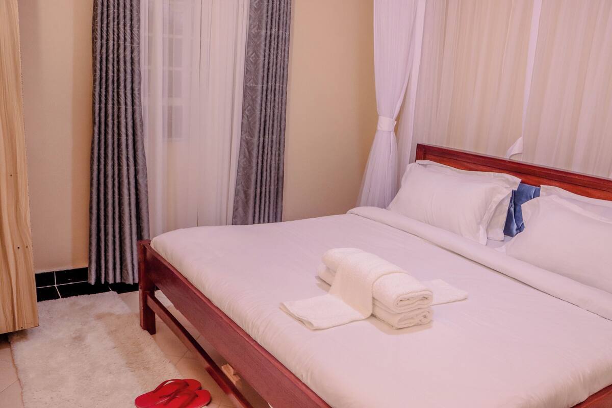 A neatly made bed with white linens and a blue pillow, a wooden headboard, and a pair of red slippers on a white rug.