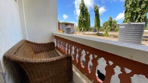 A balcony with a wicker chair, a white wall with peeling paint, and a view of trees and storage tanks. - Lux Suites Mtwapa Beach Road Apartments (Mtwapa)