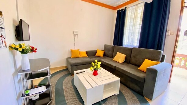 A living room with a grey sofa, yellow cushions, a white coffee table with a red vase, and a small TV on a stand. - Lux Suites Mtwapa Beach Road Apartments (Mtwapa)