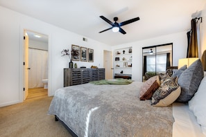 A bedroom with a bed, a ceiling fan, a dresser, a shelf with decorative items, and a window with curtains. - Clean & Modern - 4 guests - Pool & Tennis (Kill Devil Hills)