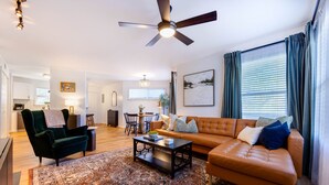 A spacious living room with a large sofa, a coffee table, and a patterned rug. There is a ceiling fan, a dining area with a table and chairs, and a kitchen in the background. - Clean & Modern - 4 guests - Pool & Tennis (Kill Devil Hills)