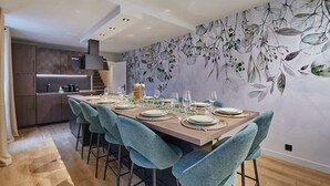 A modern dining area with a long table set for six, surrounded by teal chairs. A kitchen with dark cabinets and a built-in stove is visible in the background. - Sublime triplex Paris Le Marais (Paris)