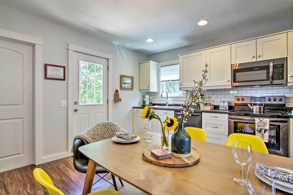 A modern kitchen with white cabinets, a built-in microwave, and a stove. There is a dining table with yellow chairs and a vase with sunflowers.
