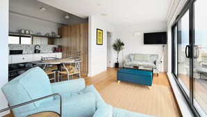 A modern kitchen with a dining area, a living room with a sofa and a TV, and a large window with a view.