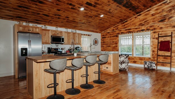A wooden cabin interior with a kitchen area featuring a stainless steel refrigerator, wooden cabinets, a microwave, and a bar with four stools. - Red River Ranch Retreat- HOT TUB, Views, Pond- Near RRG (Frenchburg)