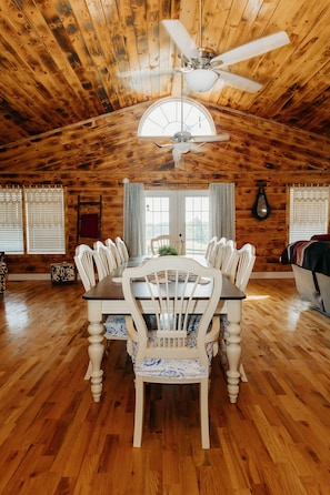 Une salle à manger en bois avec une grande table et des chaises, un ventilateur de plafond et des fenêtres avec des rideaux.