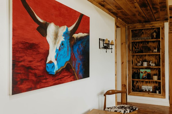 Une pièce dans laquelle on trouve, au mur, un grand tableau représentant la tête d’un taureau aux couleurs bleu et blanc, une étagère en bois sur laquelle sont disposés divers objets, et une chaise agrémentée d’un coussin à motifs.