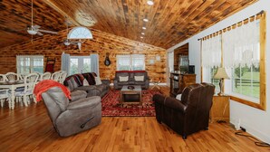 A cozy log cabin interior with a wooden ceiling, a dining area with white chairs, a sofa set, and a television. - Red River Ranch Retreat- HOT TUB, Views, Pond- Near RRG (Frenchburg)