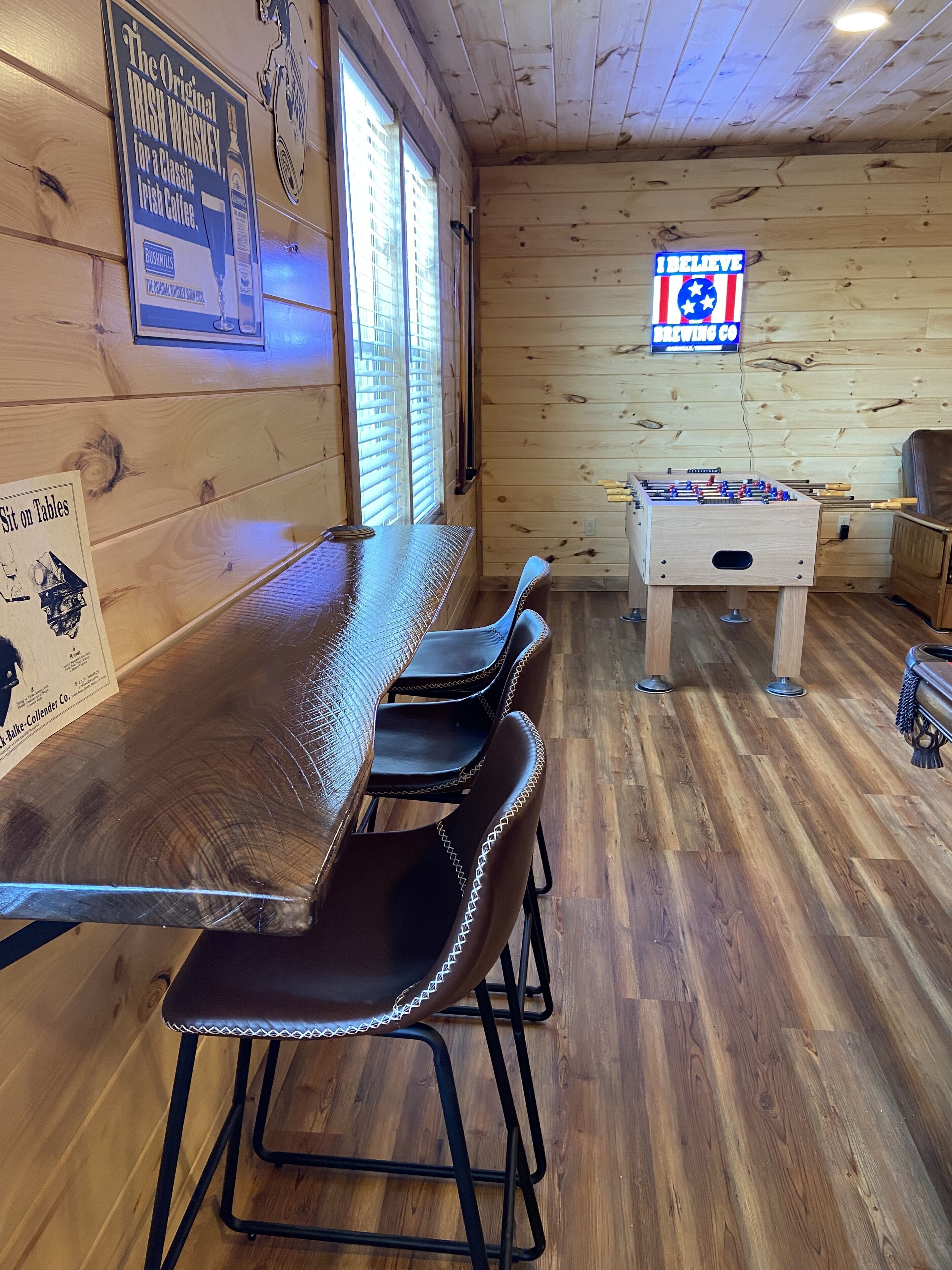 A wooden interior with a long table and four chairs, a foosball table, and a sign reading "I believe BREWING CO."