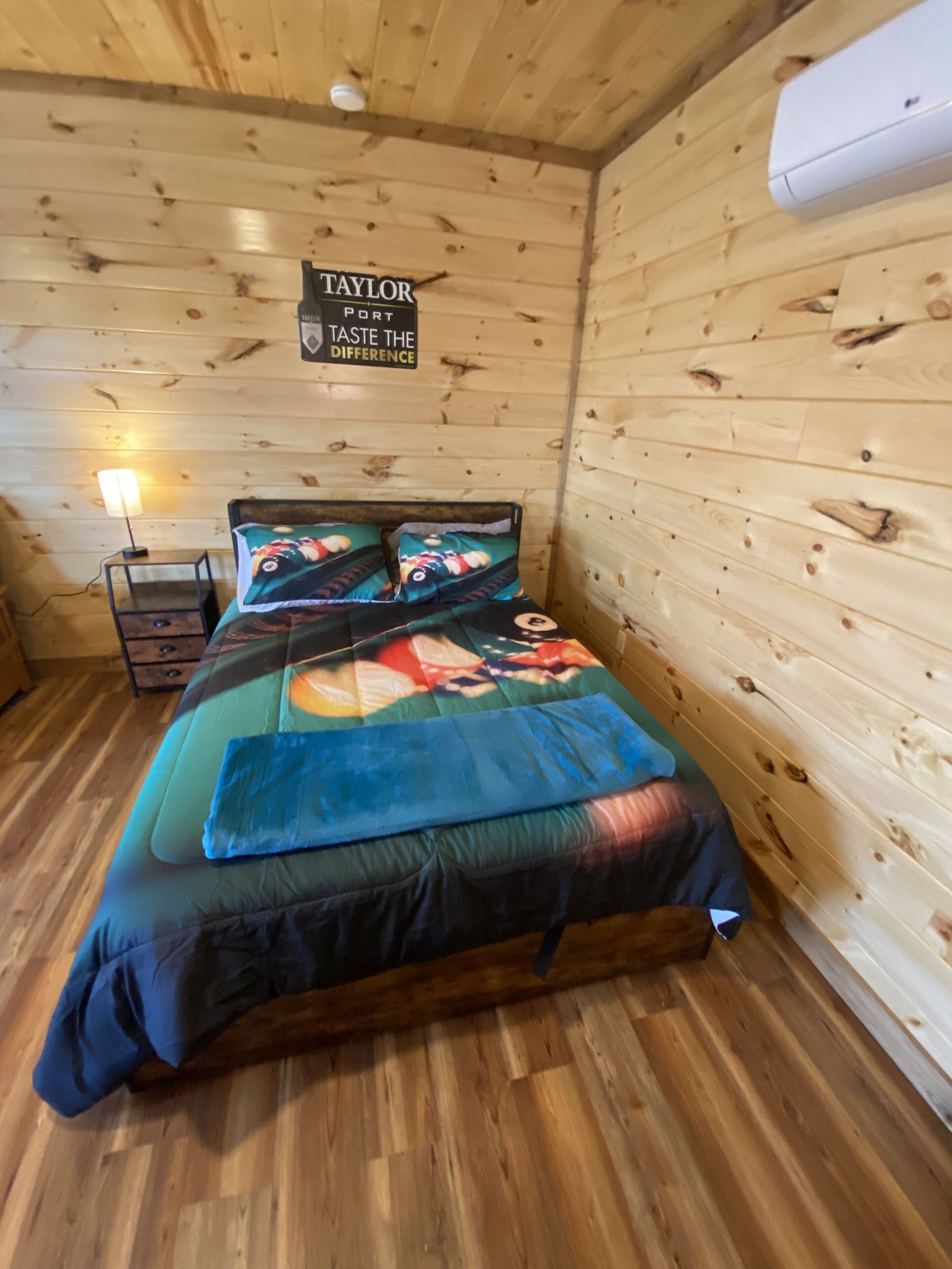 A wooden cabin room with a bed, a nightstand, and a sign that reads "TAYLOR PORT TASTE THE DIFFERENCE".