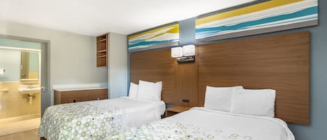 Quarto de hotel com duas camas, uma cabeceira de madeira, banheiro com pia e espelho, e parede com arte abstrata.