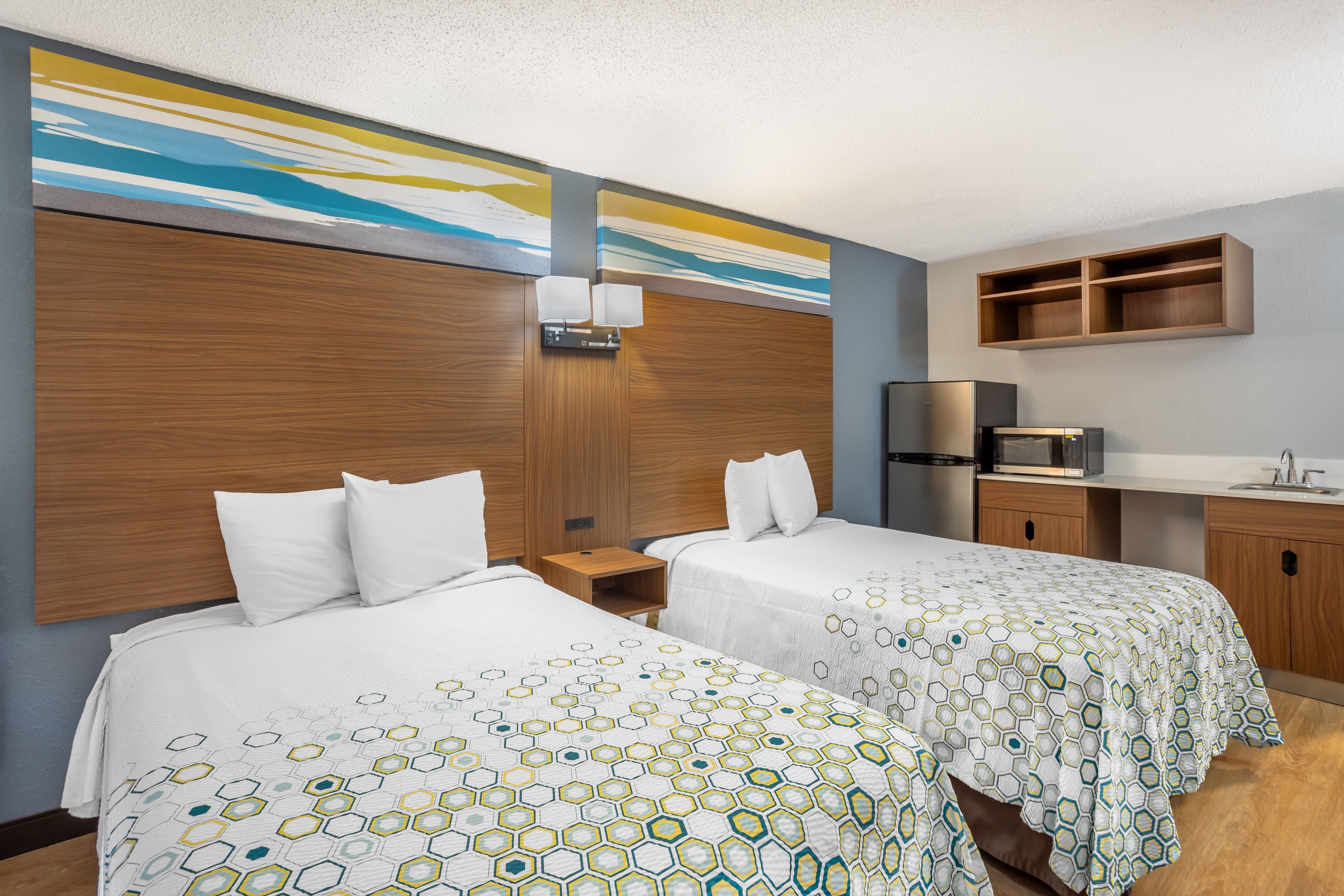 A hotel room with two beds, a wooden headboard, a small desk, a refrigerator, and a built-in shelf above the counter.