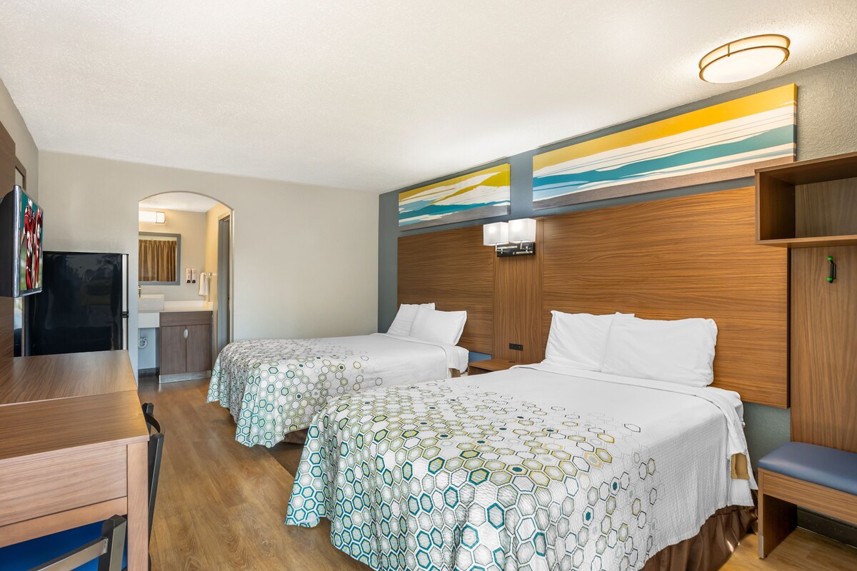 Habitación de hotel con dos camas, un escritorio y un televisor. Se ve un baño a través de un arco.