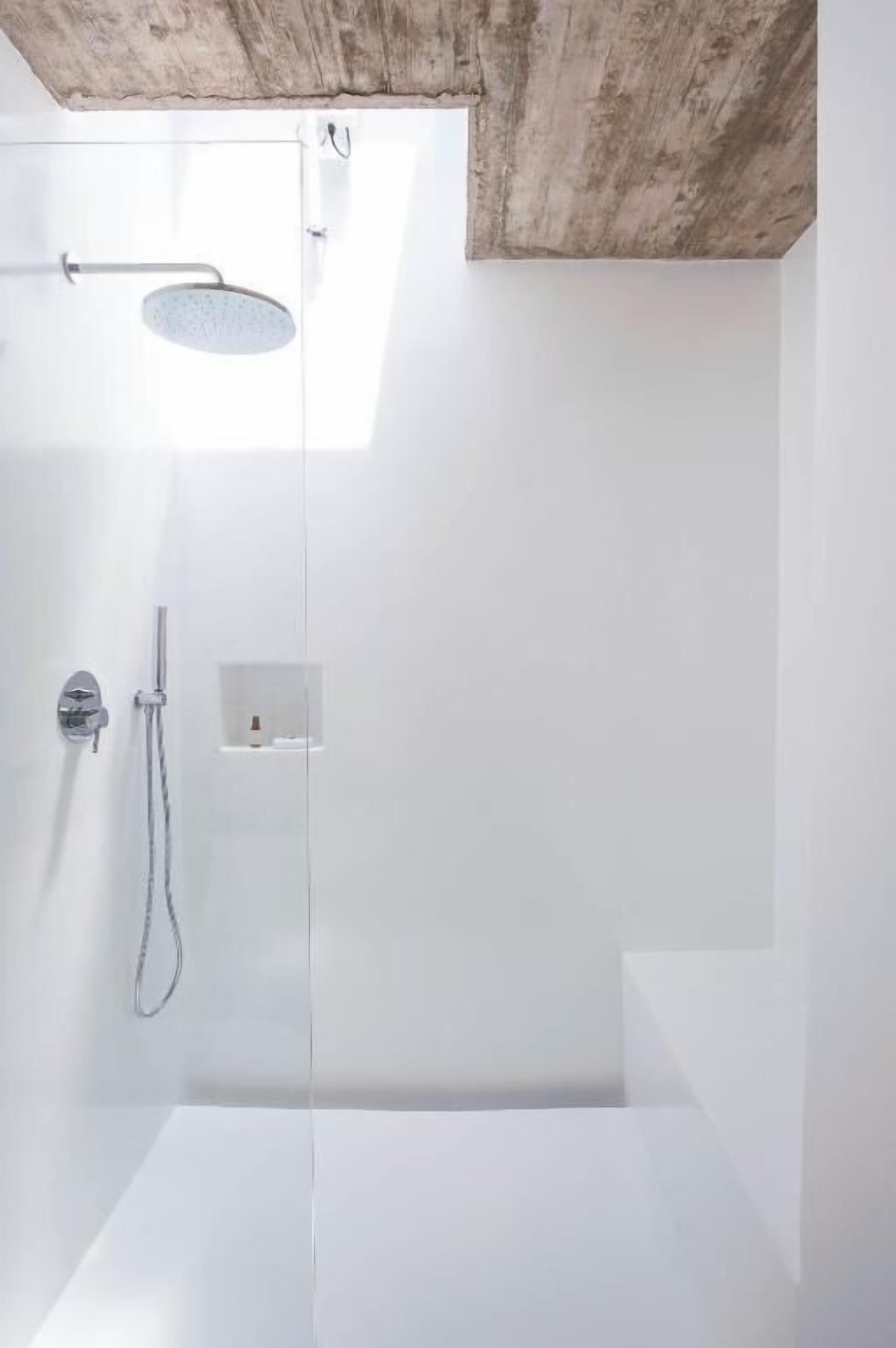 A modern shower area with a rain showerhead and handheld showerhead, a small shelf with toiletries, and a concrete ceiling.