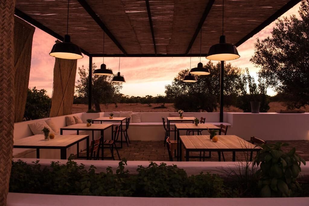 Un área de estar al aire libre con mesas y bancos bajo una pérgola de madera, rodeada de vegetación y árboles.