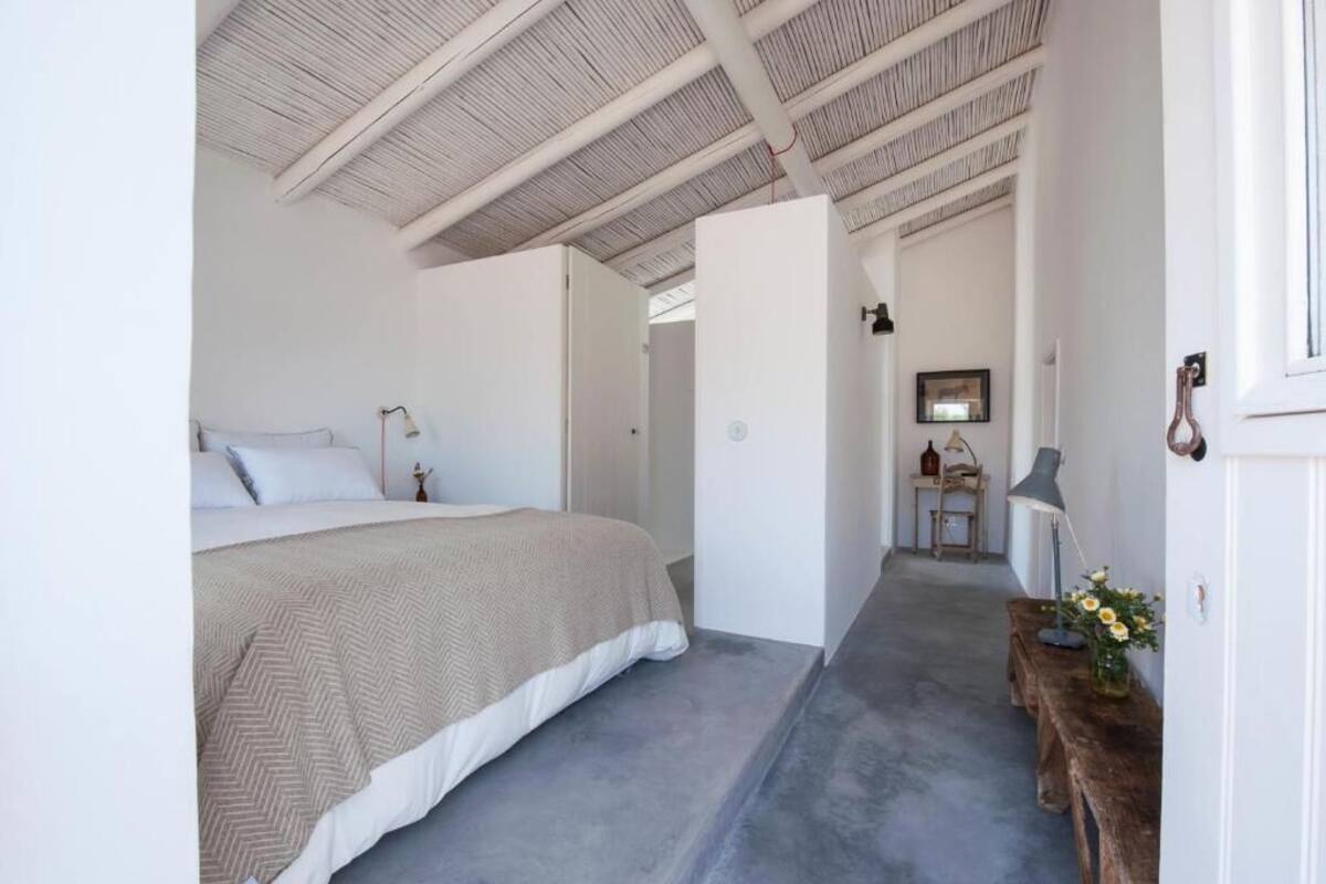 A bedroom with a large bed, a wooden bench with flowers, a desk with a lamp, and a mirror on the wall.