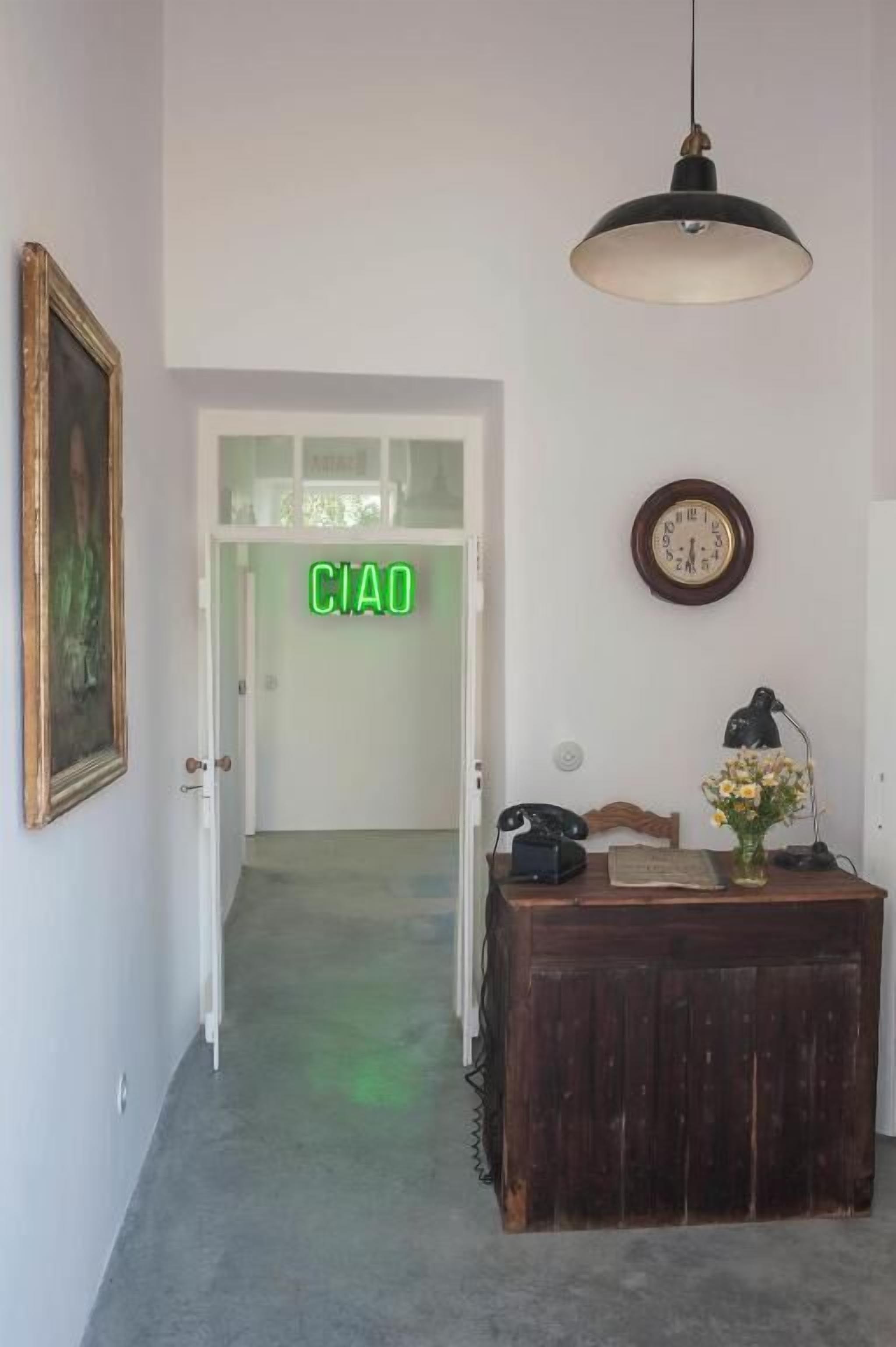 A hallway with a wooden door labeled "CIAO," a desk with a telephone, a lamp, and a vase of flowers.