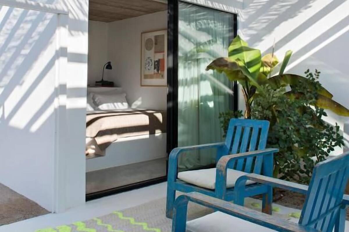 A modern outdoor seating area with a blue chair and a green rug, a glass door leading to a bedroom with a bed and a nightstand, and a large plant in the background.