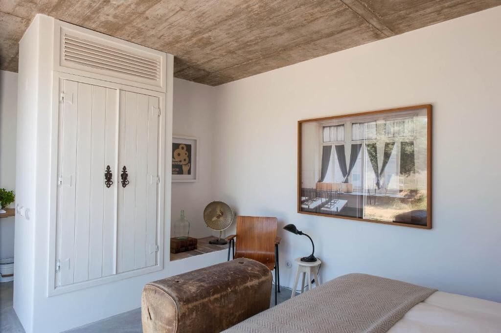 Ein Schlafzimmer mit großem Kleiderschrank, einem Bett, einem kleinen Tisch mit Lampe, einem Sessel und einem gerahmten Bild an der Wand.