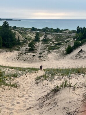 A person standing on a sandy path with dunes and sparse vegetation, overlooking a body of water and distant land.