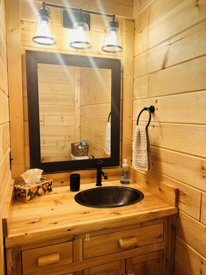 A wooden cabin-style bathroom with a large mirror, three pendant lights, a black sink, a towel hanging on a hook, a tissue box, and a soap dispenser.