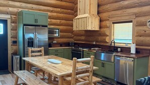 A log cabin interior with a wooden dining table, chairs, a kitchen with modern appliances, and a large window.