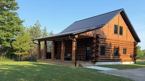 A log cabin with a covered porch, a gravel driveway, and a grassy yard.