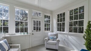 Una habitación luminosa con ventanas amplias, una puerta blanca, un sillón acolchado y un banco con cojines estampados.
