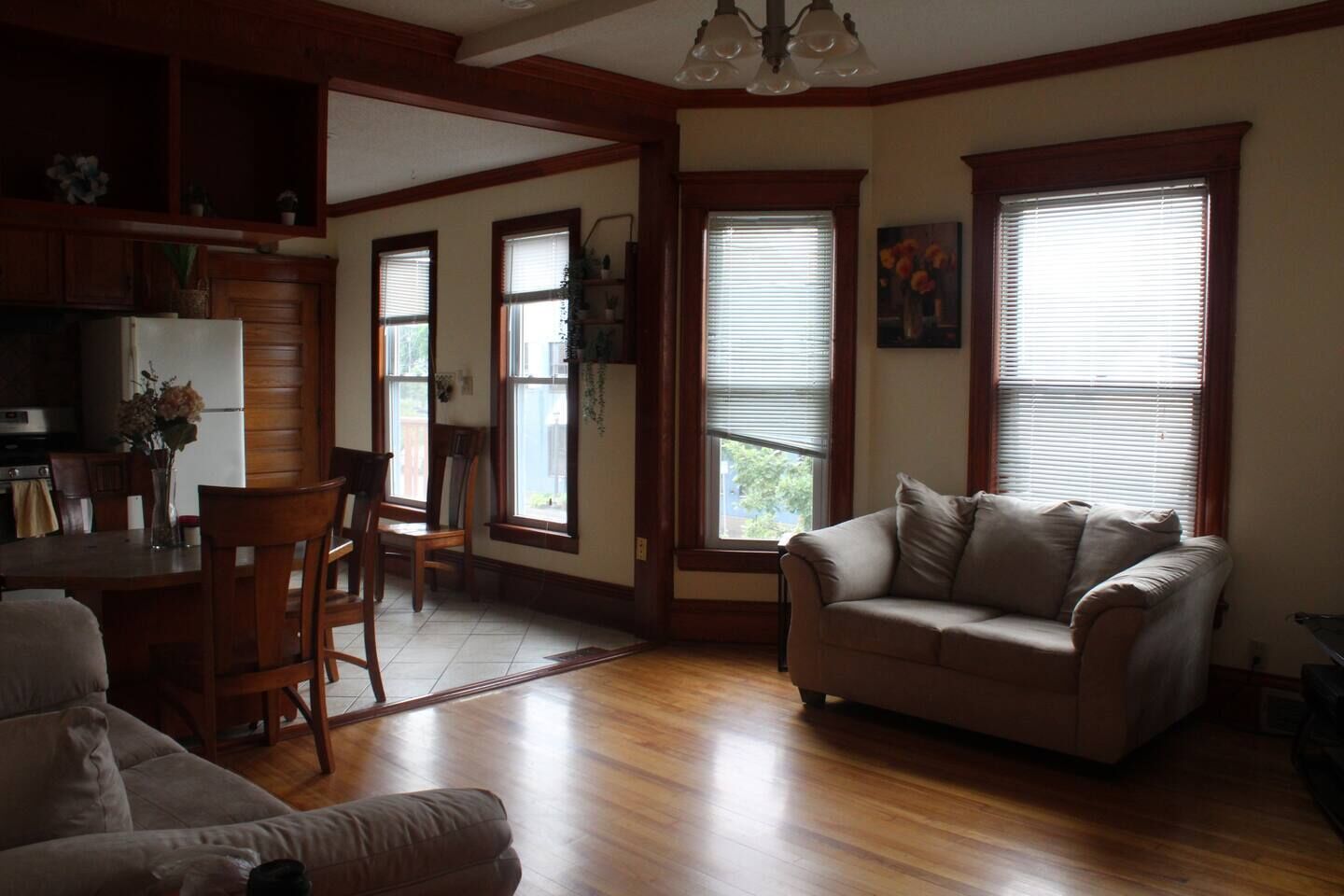 A spacious living room with a beige sofa, a dining table with chairs, and a kitchen area with wooden cabinets.