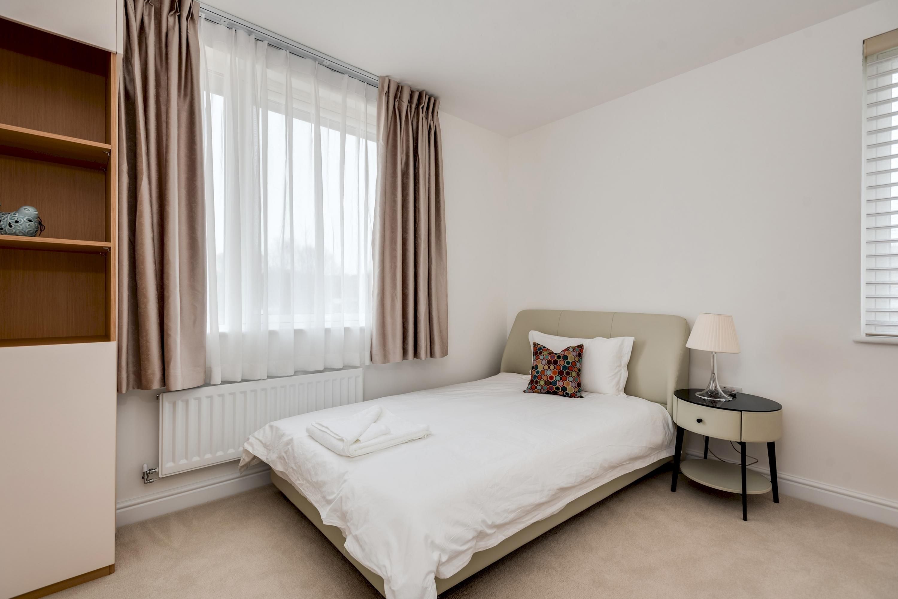 A neatly made bed with a white duvet, a small bedside table with a lamp, and a window with curtains.