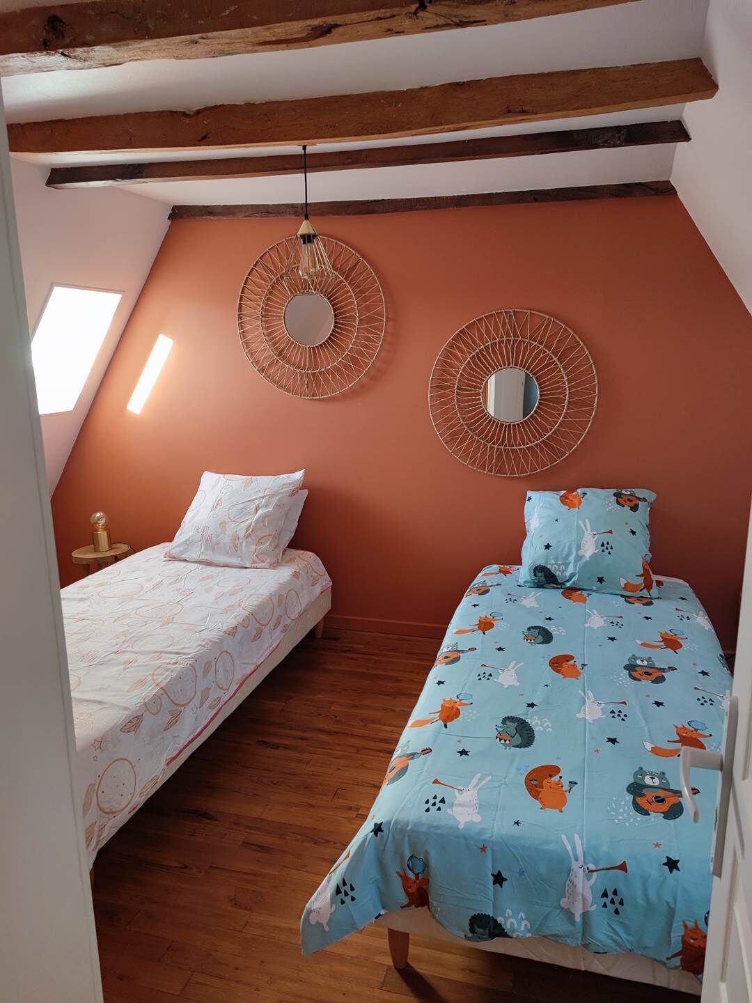 A bedroom with two beds, one with a white bedspread and the other with a blue bedspread featuring animal prints. There are two wall-mounted mirrors and a skylight above the beds.
