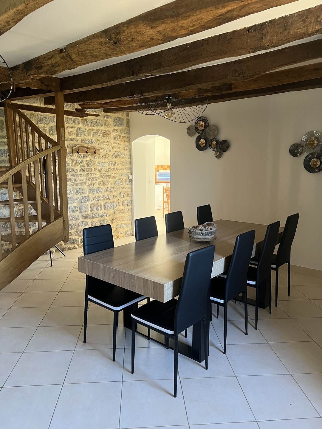 A dining area with a large wooden table, six black chairs, a stone wall, and a wooden staircase.