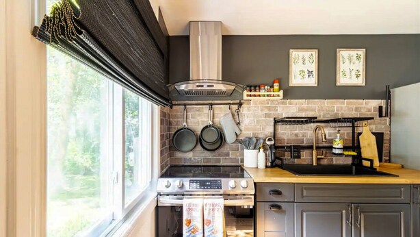 Cocina compacta con estufa, horno y ollas colgantes. Hay dos láminas botánicas enmarcadas en la pared y una ventana con persianas.