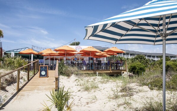 Un restaurante frente al mar con terraza al aire libre bajo sombrillas a rayas, una cubierta de madera que conduce a la entrada y un letrero dando la bienvenida a los huéspedes.