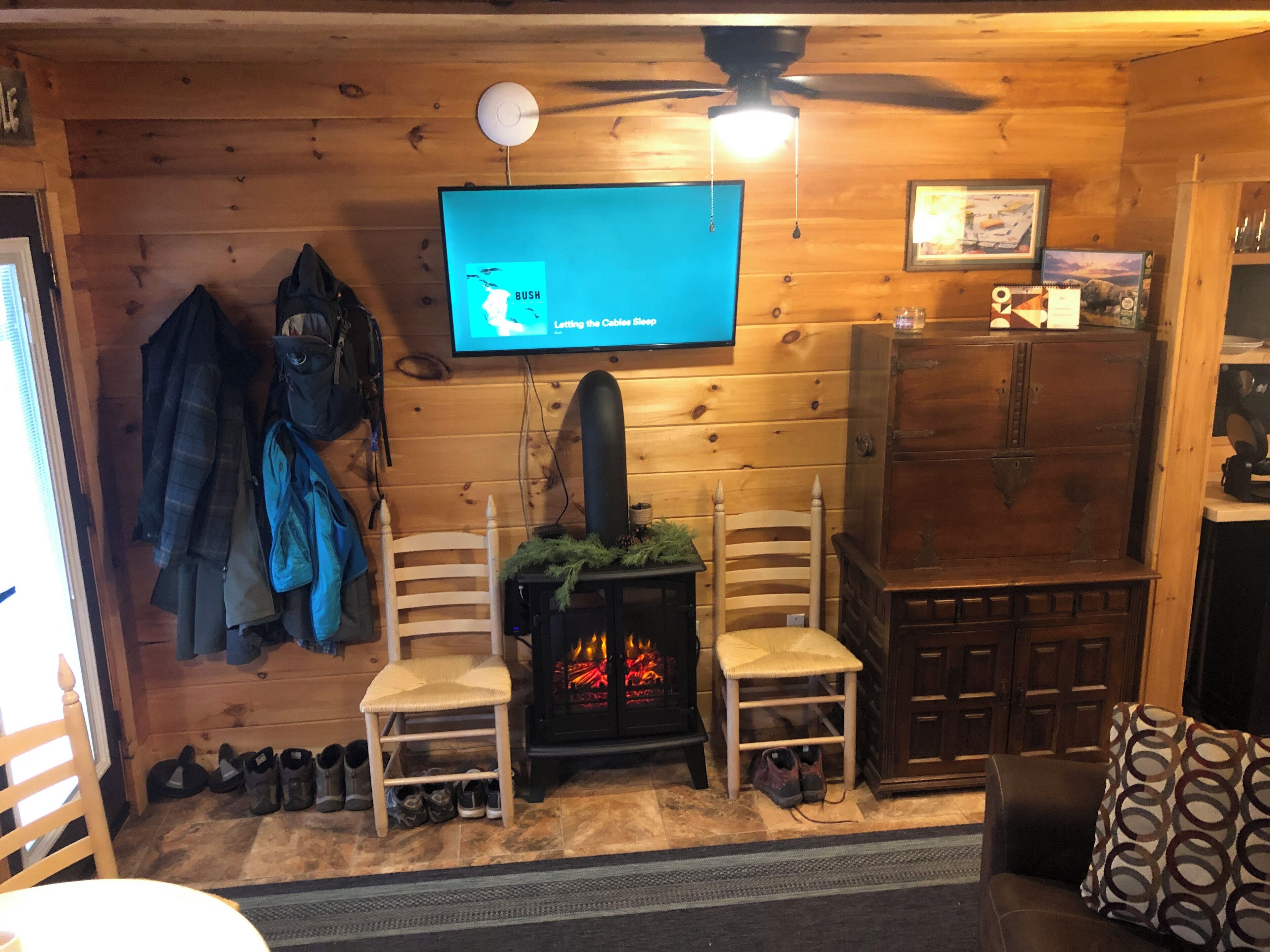 Een gezellig chaletinterieur met een open haard, twee stoelen, een aan de muur bevestigde televisie en een houten kast.