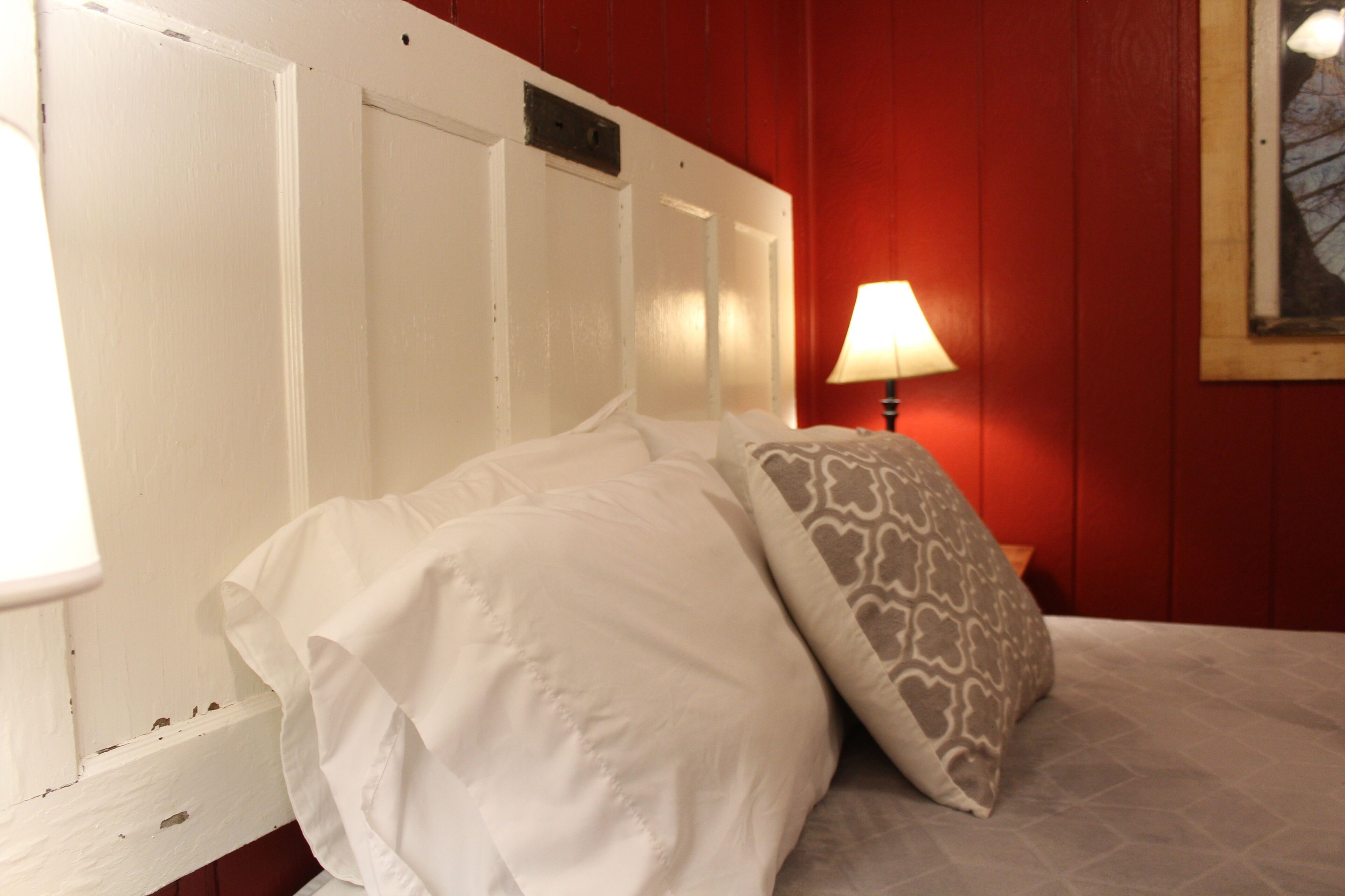 A bed with white bedding and a patterned pillow in a room with red walls and a white door.