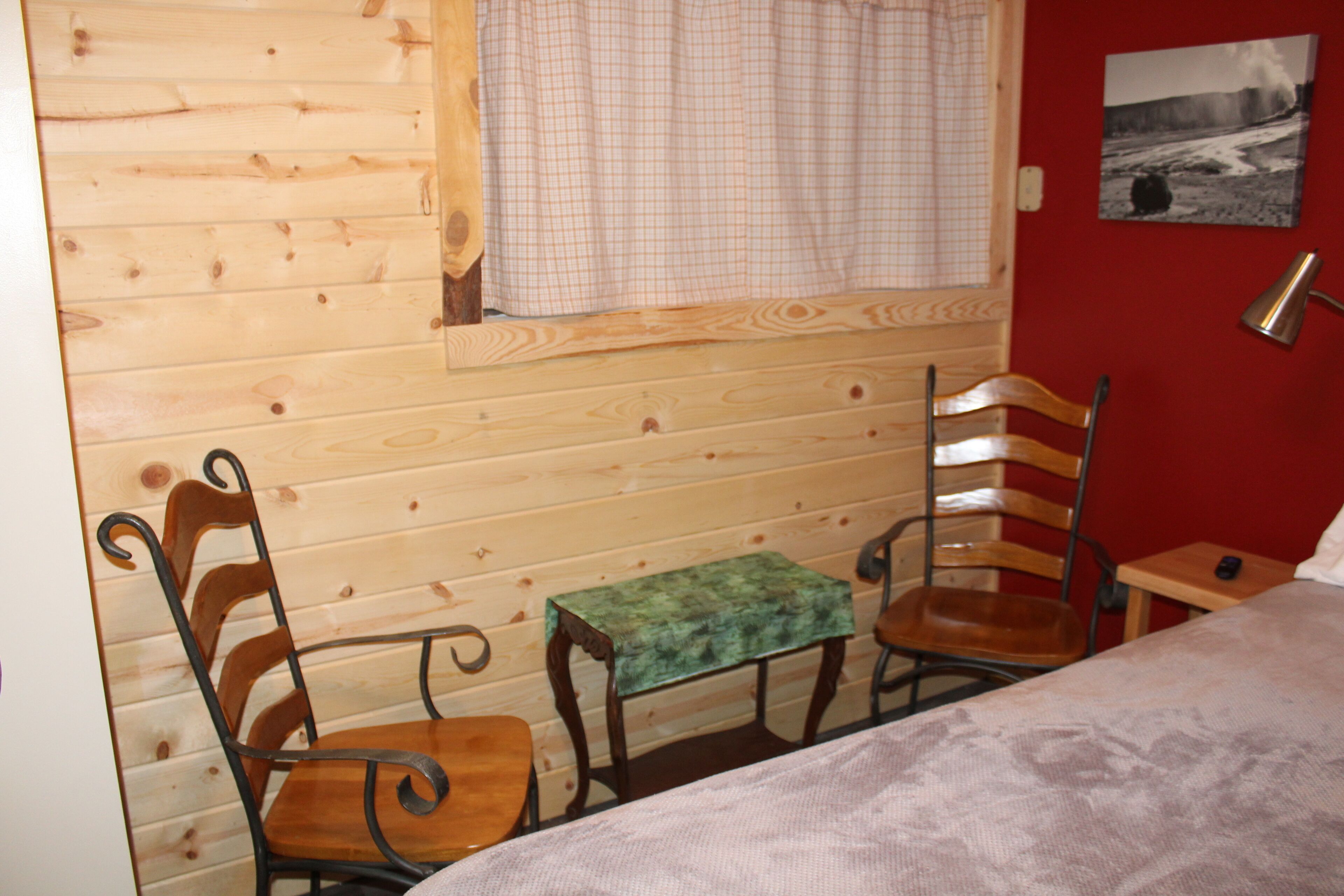 Habitación de cabaña de madera con una cama, dos sillas de madera, una mesita y una pared roja con un cuadro blanco y negro.