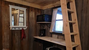 A compact kitchen with wooden walls, a microwave, a coffee maker, a small refrigerator, a wooden ladder, and a window with curtains.