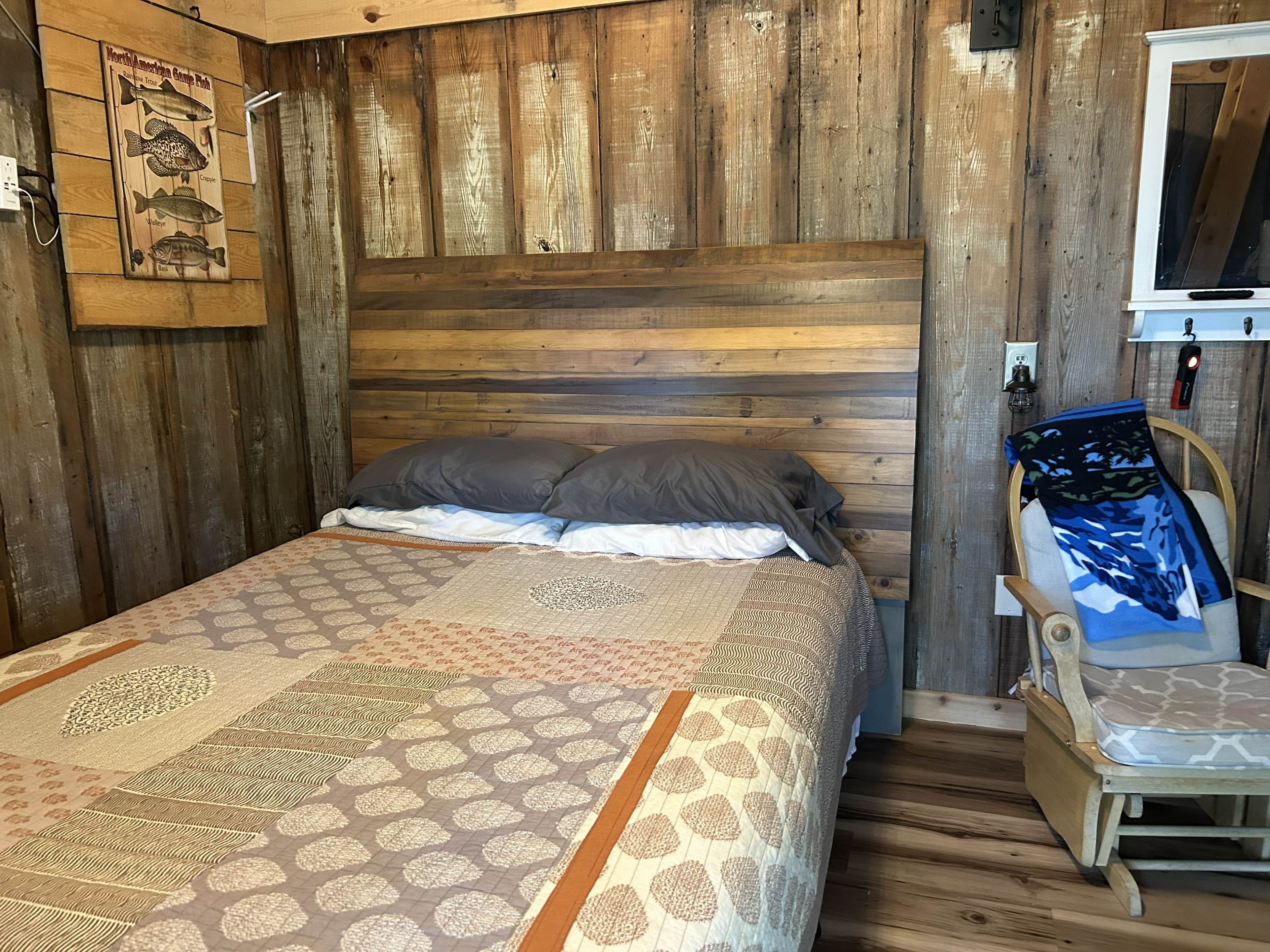 A wooden-paneled room with a bed, pillows, a patterned bedspread, a chair, and a wall-mounted TV.