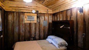 A wooden cabin room with a bed, a ceiling fan, a wall-mounted heater, and a small window.