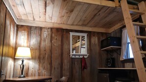 A rustic wooden room with a table, chairs, a lamp, a window with curtains, a shelf with a microwave, and a ladder.