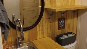 A bathroom with a wooden vanity, a stainless steel sink, a mirror, and a small shelf with toiletries.