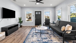 A modern living room with a flat-screen TV, a ceiling fan, a black sofa, a coffee table, and a large window. - Inviting 4br/2.5ba Downtown Home With Ample Space! (San Antonio)