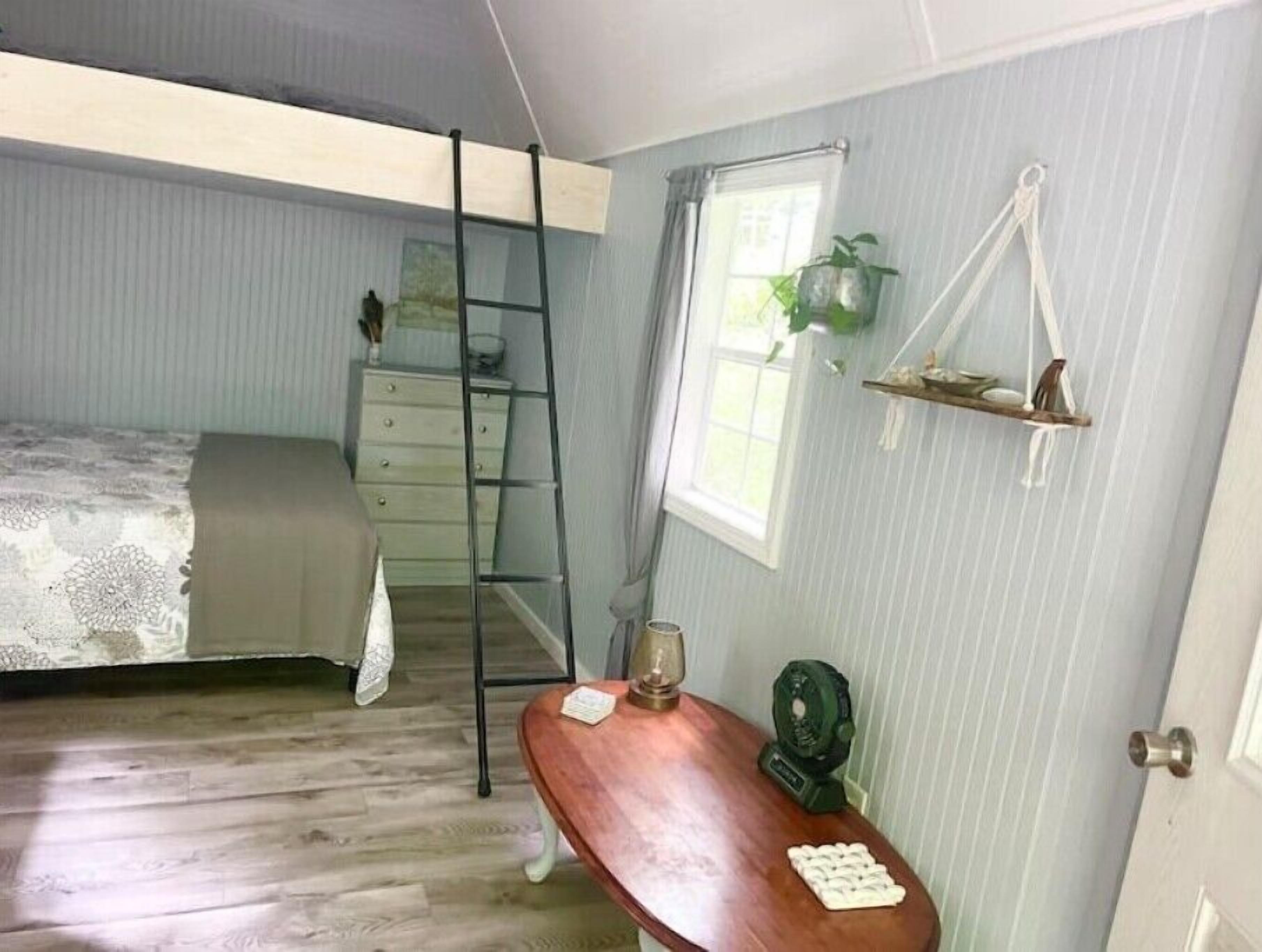Ein Schlafzimmer mit einem Bett, einer Leiter, einem Schminktisch, einem kleinen Tisch mit einer Pflanze und einem Fenster.